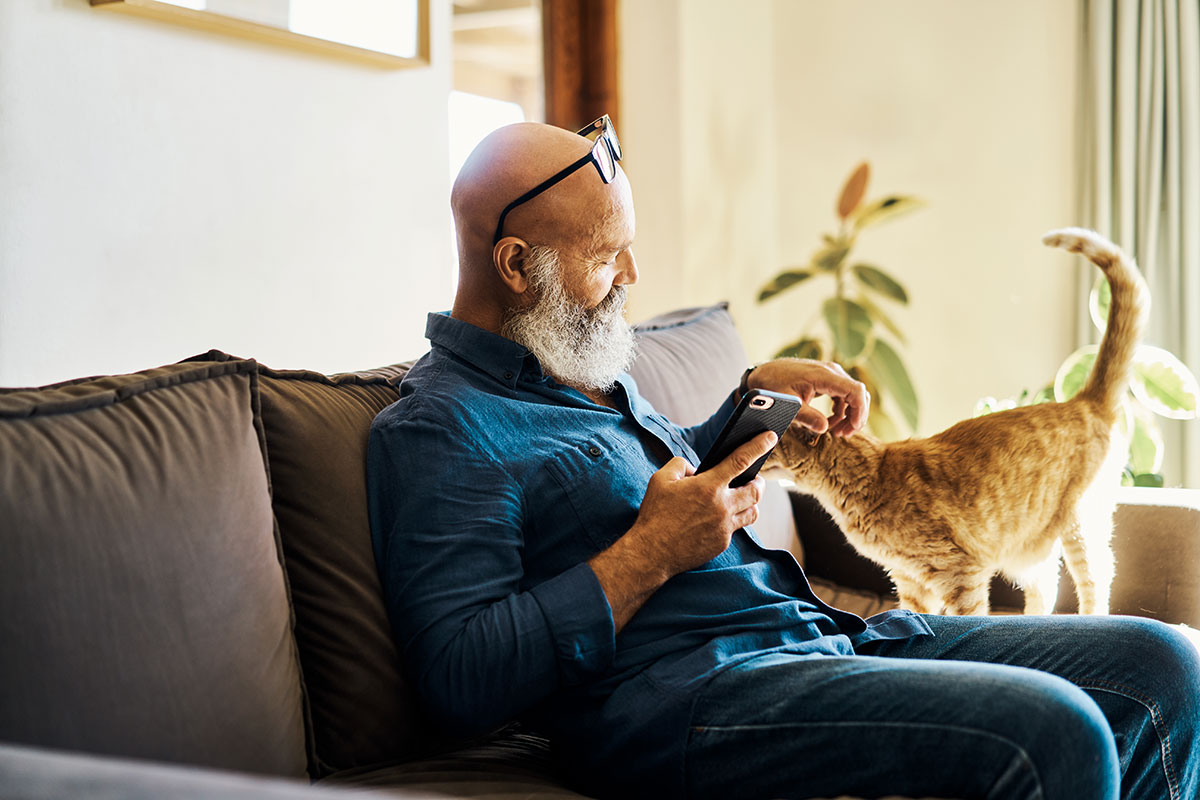 man sitting on couch petting a cat and holding his mobile phone